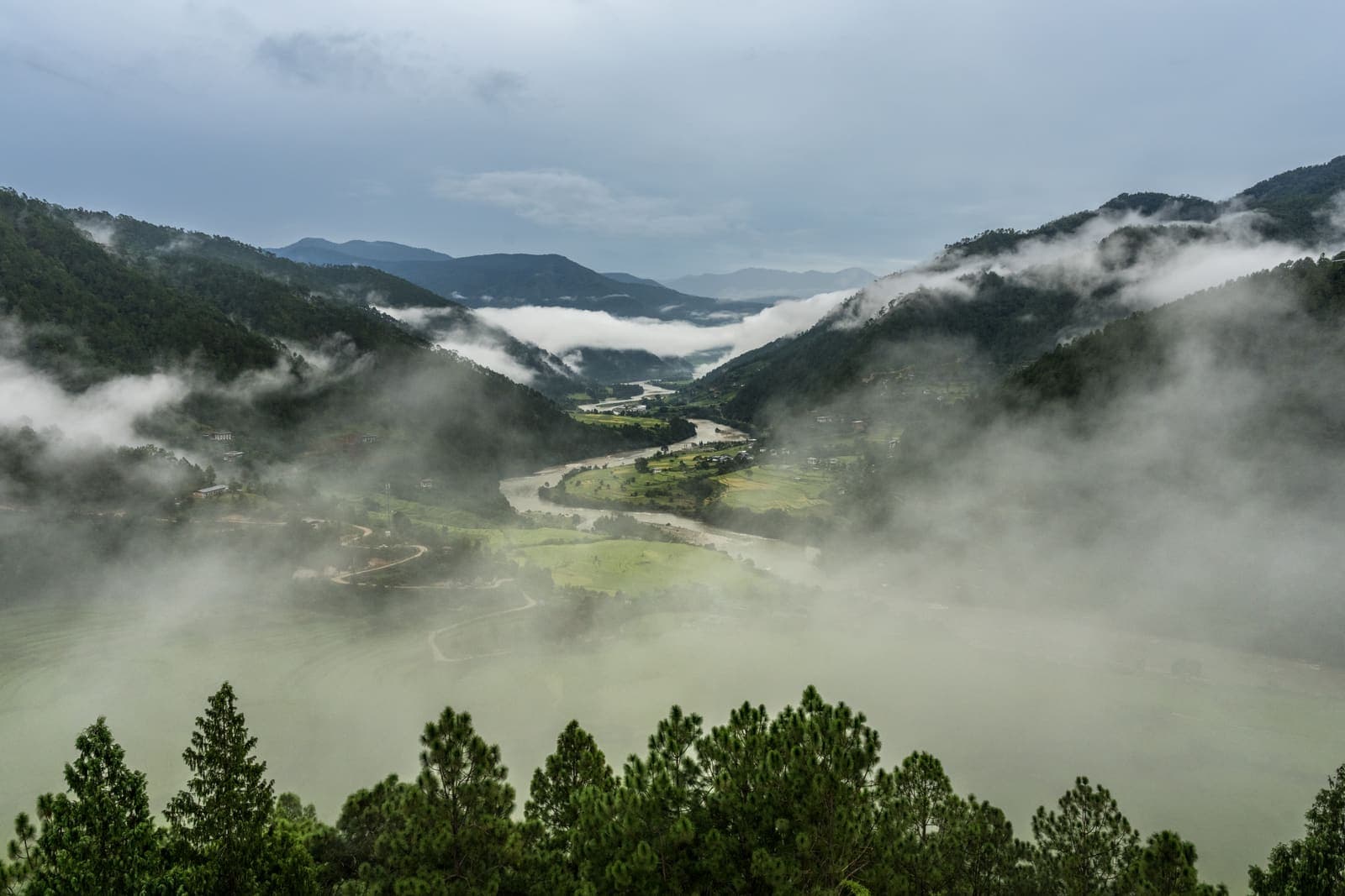 A green Bhutanese river valley with cloud drifting through the forested hills